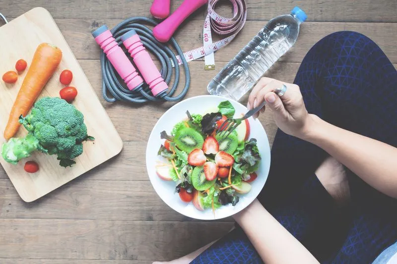 mulher praticando hábitos saudáveis, segurando um prato de salada saudável, e na mesa ao fundo uma garrafa de água, halteres e corda de pular.
